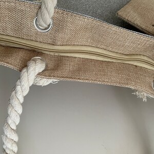 May include: A beige canvas tote bag with a zipper closure and rope handles. The bag has a small pocket on the outside.