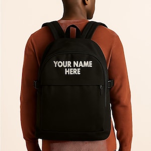 May include: Black backpack with the text "YOUR NAME HERE" in white. The backpack has adjustable straps and a front pocket. The person in the image is wearing a rust-colored sweater.