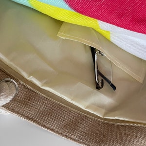 May include: A beige canvas tote bag with a zipper closure and a rope handle. The bag has a light beige lining with a zippered pocket inside. A pair of black glasses is inside the pocket.