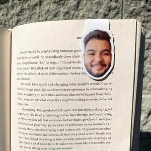 May include: A white bookmark with a smiling man's face on it is placed on the pages of a book. The bookmark is in the shape of the man's face. The book is open, and the text is visible.