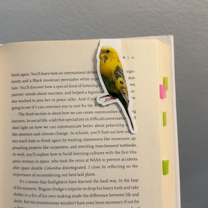 May include: A yellow and green parakeet sticker is placed on the edge of a book. The sticker has a white border and is in the shape of a bird. The book is open to a page of text with colorful sticky notes.