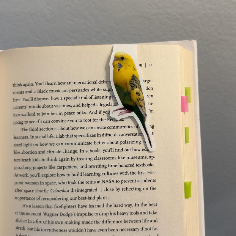 May include: A yellow and green parakeet sticker is placed on the edge of a book. The sticker has a white border and is in the shape of a bird. The book is open to a page of text with colorful sticky notes.