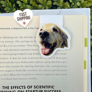 May include: A bookmark featuring a smiling golden retriever dog. The bookmark is white with a die-cut shape of the dog's head. The image is on a book with the text "FAST SHIPPING" on a heart-shaped sticker.