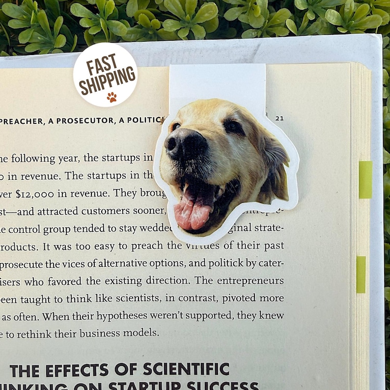 May include: A bookmark featuring a smiling golden retriever dog face. The bookmark is white with a die-cut shape and is placed on the pages of a book. A round sticker with the text "FAST SHIPPING" is also visible.