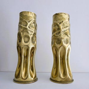 May include: A pair of brass vases. The upper section has a textured, leaf-like pattern, while the lower part features a fluted, curved design. The vases have a shiny, golden finish and a flared base. They are set against a white backdrop.