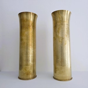 May include: Two tall, cylindrical brass vases with a textured surface. The vases have a flared top and a simple, rounded base. The brass has a warm, golden hue, and the vases are displayed against a white background.