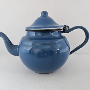 Op de afbeelding: Een blauwe emaille theepot met een afgeronde body, gebogen tuit en bijpassende deksel. De theepot heeft een zwarte rand en handvat. Het handvat is gebogen en aan de zijkant van de theepot bevestigd. De theepot is een klassiek ontwerp.