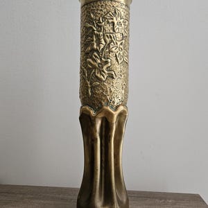May include: A gold metal vase with a floral design. The vase is made from a repurposed shell casing and has a flared base.