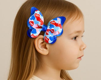 Puede incluir: Una pinza para el pelo con forma de mariposa azul con patrones de corazones rojos, blancos y azules y la palabra "USA". El accesorio para el pelo se lleva en el lateral de la cabeza de una niña.
