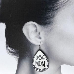 Puede incluir: Un par de pendientes colgantes con un dise&ntilde;o en forma de l&aacute;grima. Los pendientes est&aacute;n hechos de piel sint&eacute;tica negra y blanca con un patr&oacute;n de huellas de patas y huesos. Los pendientes tienen un gancho plateado y el texto "DOG MOM" est&aacute; impreso en blanco en la parte delantera.