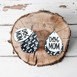 Puede incluir: Un par de pendientes colgantes con un patr&oacute;n de huellas de patas y huesos en blanco y negro. Un pendiente tiene un &oacute;valo blanco con el texto "Dog Mom" en negro. El otro pendiente tiene un &oacute;valo negro con el texto "Dog Mom" en blanco.