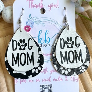 Puede incluir: Un par de pendientes colgantes con un dise&ntilde;o de huella de pata en blanco y negro. Los pendientes est&aacute;n hechos de cuero y tienen forma de l&aacute;grima. El texto "DOG MOM" est&aacute; impreso en los pendientes.