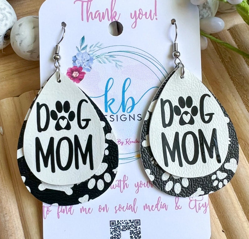 Puede incluir: Un par de pendientes colgantes con un dise&ntilde;o de huella de pata en blanco y negro. Los pendientes est&aacute;n hechos de cuero y tienen forma de l&aacute;grima. El texto "DOG MOM" est&aacute; impreso en los pendientes.
