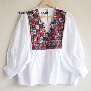 May include: White blouse with a colourful embroidered yoke. The yoke features a geometric pattern with red, blue, and pink squares, and small mirrors. The long-sleeved top is hanging on a wooden hanger.