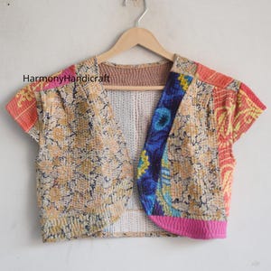 Puede incluir: Bolero patchwork con mangas cortas, con una mezcla de telas estampadas florales y otros diseños en tonos amarillos, naranjas, azules y rosas. La chaqueta está colgada de una percha de madera sobre un fondo blanco.