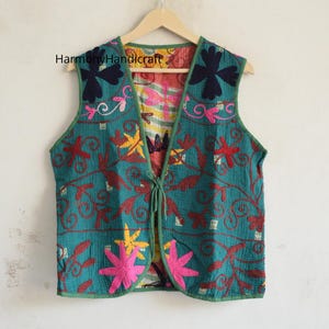May include: A sleeveless waistcoat in teal fabric featuring intricate floral embroidery in pink, yellow, and dark blue. The garment has a front tie closure and green trim. The text HarmonyHandicraft is visible on the upper left side of the waistcoat.