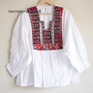 Può includere: Top in cotone bianco con carrè ricamato dettagliato in rosso, nero e argento. Il top ha maniche lunghe e una vestibilità ampia. Il testo "HarmonyHandicraft" è visibile in alto a sinistra.