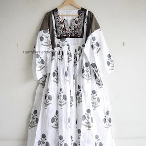 Puede incluir: Vestido blanco con estampado floral y canes&uacute; marr&oacute;n. El vestido tiene mangas largas y un escote en V con detalles bordados. El vestido est&aacute; colgado de una percha de madera.