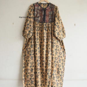 Puede incluir: Un vestido largo y fluido con un estampado floral en tonos beige y azul. El vestido presenta un canesú detallado con un diseño rojo oscuro y negro. Las mangas son de tres cuartos y el vestido cuelga de una percha de madera.