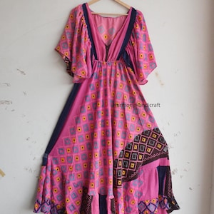 Może przedstawiać: Różowa sukienka we wzory z dekoltem w serek i krótkimi, rozkloszowanymi rękawami. Sukienka ma geometryczny wzór w kolorze żółtym, niebieskim i różowym. Granatowe wykończenia podkreślają dekolt i boki. Sukienka wisi na drewnianym wieszaku.