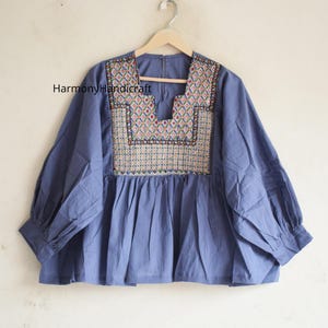 May include: A blue blouse with long sleeves and a square neckline featuring intricate multi-colored geometric embroidery on the chest. The garment has a gathered waistline and includes the text HarmonyHandicraft printed on the upper left side.