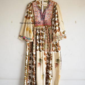 May include: A long, cream-colored dress with a colorful embroidered neckline and paisley and floral patterns. The dress has long, wide sleeves and is hanging on a wooden hanger. The text "Harmony Handicraft" is visible.