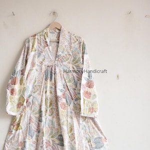 Puede incluir: Un vestido largo blanco con un estampado floral en tonos rosa, verde y azul cuelga de una percha de madera. El vestido tiene mangas largas y un escote en V. El texto "HarmonyHandicraft" es visible en el vestido.