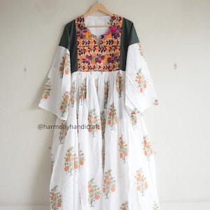 May include: A white dress with orange floral print and a dark green yoke. The yoke features colourful embroidery and a V-neck. The dress has wide sleeves and is hanging on a wooden hanger.