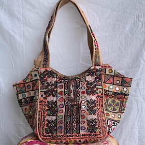 Könnte beinhalten: Eine Umhängetasche mit Patchwork-Design, aufwendigen mehrfarbigen Stickereien, kleinen runden Spiegeln und geometrischen Mustern. Die Tasche hat eine breite Öffnung, einen einzelnen Schulterriemen und besteht aus verschiedenen Stoffbahnen.