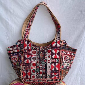 Puede incluir: Un bolso tipo tote con intrincados bordados multicolores en patrones geométricos rojos, negros y blancos. El bolso está adornado con pequeños espejos circulares en todo el diseño e incluye dos correas de tela color canela.