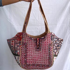 May include: A tote bag featuring a patchwork design with vibrant pink, red, and multi-colored embroidered fabric panels. The bag includes small circular mirror accents, intricate geometric stitching, and two tan shoulder straps for carrying.