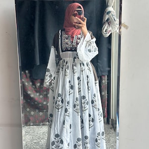 Puede incluir: Un vestido largo blanco con un estampado floral en negro y gris. El vestido tiene mangas largas con un corpi&ntilde;o blanco y marr&oacute;n. La persona de la imagen lleva un velo rosa.