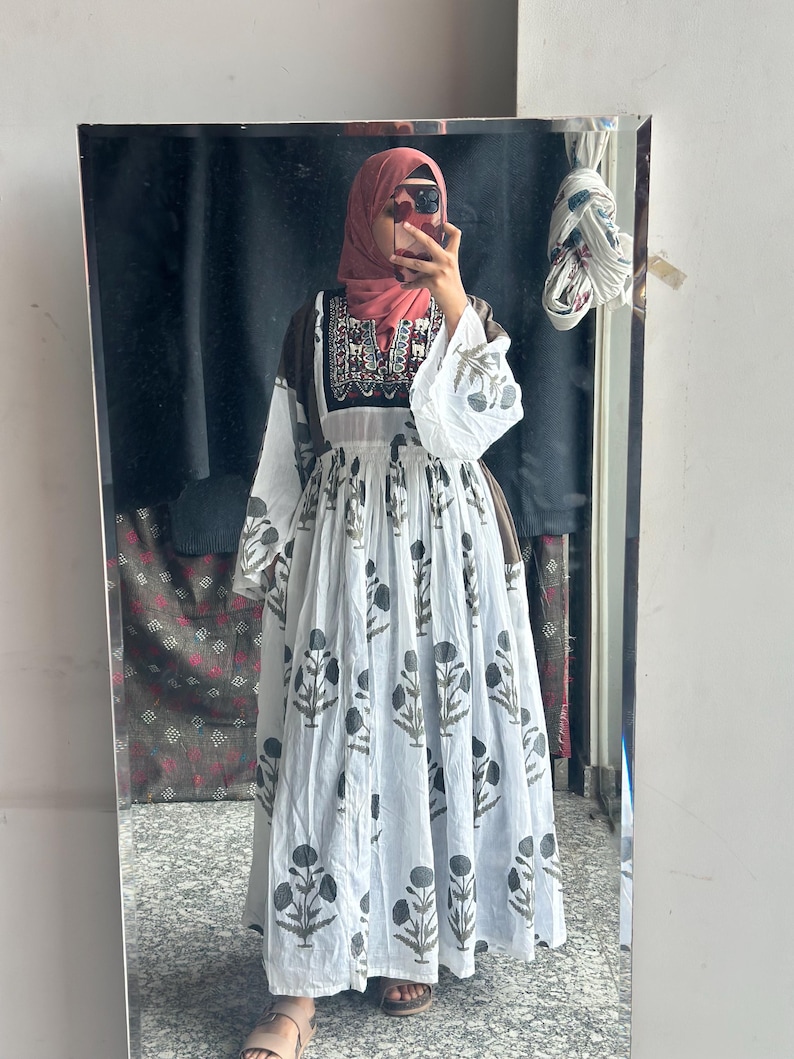 Puede incluir: Un vestido largo blanco con un estampado floral en negro y gris. El vestido tiene mangas largas con un corpi&ntilde;o blanco y marr&oacute;n. La persona de la imagen lleva un velo rosa.