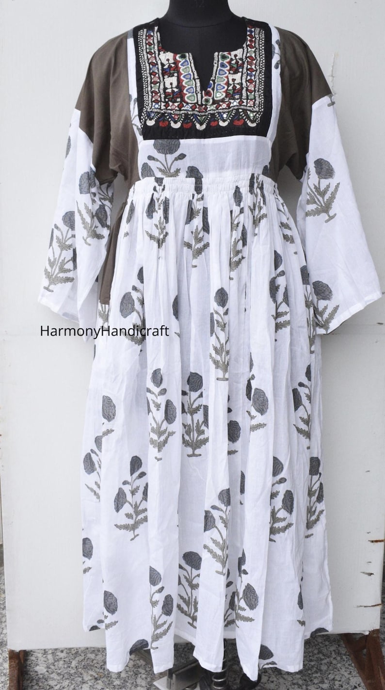Puede incluir: Un vestido largo blanco con estampado floral y mangas largas. El vestido presenta un canes&uacute; marr&oacute;n con detalles bordados y el texto "HarmonyHandicraft". El vestido se exhibe en un maniqu&iacute;.
