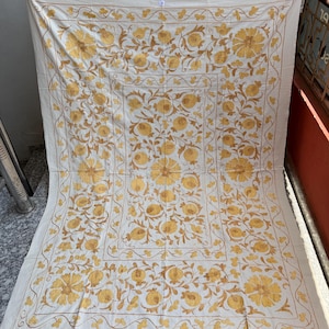 May include: Cream-colored textile with intricate embroidery. The design features large yellow floral patterns and smaller floral and vine motifs. The embroidery is in shades of yellow and brown, creating a detailed and ornate appearance. The textile is likely a decorative item.
