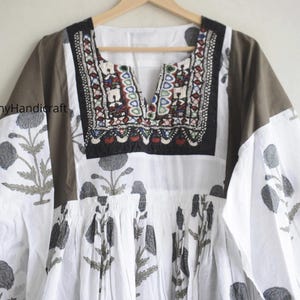 Puede incluir: Una t&uacute;nica blanca y marr&oacute;n con estampado floral y escote bordado. La t&uacute;nica presenta un cuello en V y mangas largas. El estampado floral es en tonos grises. El escote est&aacute; decorado con bordados coloridos.