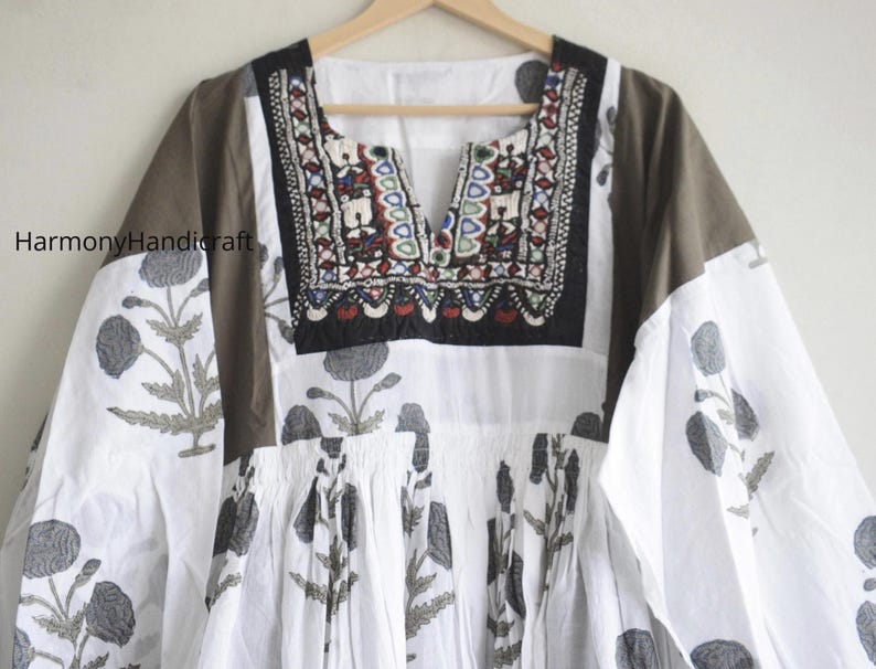 Puede incluir: Una t&uacute;nica blanca y marr&oacute;n con estampado floral y escote bordado. La t&uacute;nica presenta un cuello en V y mangas largas. El estampado floral es en tonos grises. El escote est&aacute; decorado con bordados coloridos.
