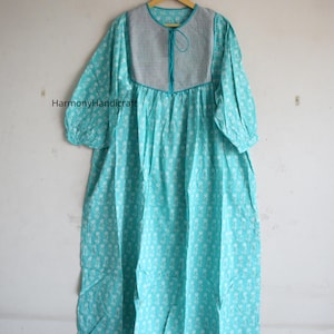 May include: A turquoise cotton dress with a white floral pattern, hanging on a wooden hanger. The dress features a gray yoke with a drawstring neckline and three-quarter sleeves. The dress is a loose, flowing style.
