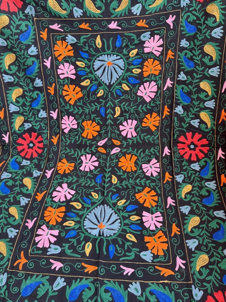 May include: Black textile with colorful floral embroidery. The design features a central floral motif with blue, pink, orange, and red flowers, surrounded by green leaves and vines. The border has a repeating paisley pattern.