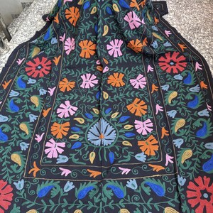 May include: Black textile with colorful floral embroidery. The design features large flowers in shades of blue, orange, pink, and red, surrounded by green leaves and vines. The edges are bordered with a repeating pattern.