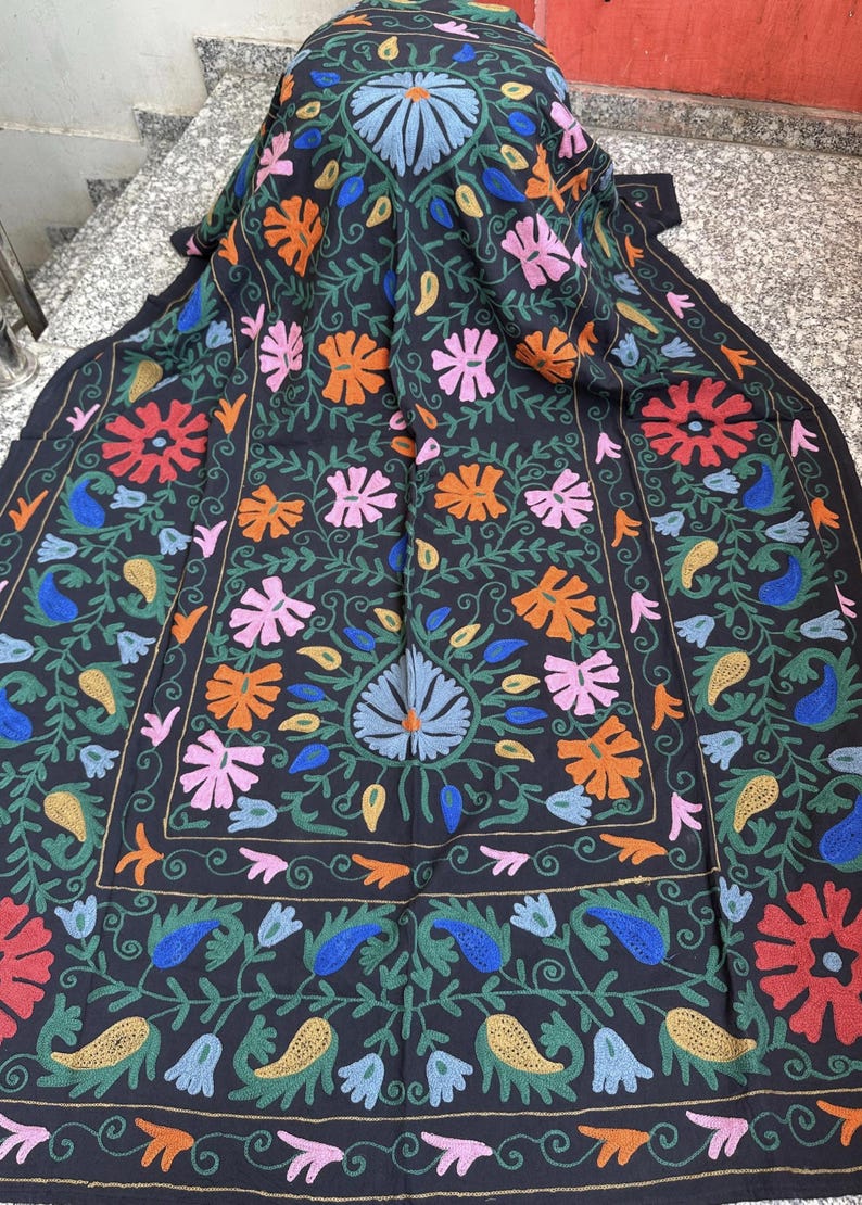 May include: Black textile with colorful floral embroidery. The design features large flowers in shades of blue, orange, pink, and red, surrounded by green leaves and vines. The edges are bordered with a repeating pattern.