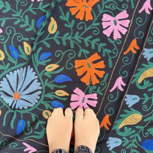 May include: Dark navy fabric with colorful floral embroidery. The design features orange, pink, blue, and yellow flowers with green vines. The embroidery is detailed and covers the fabric. The image shows a close-up of the fabric.