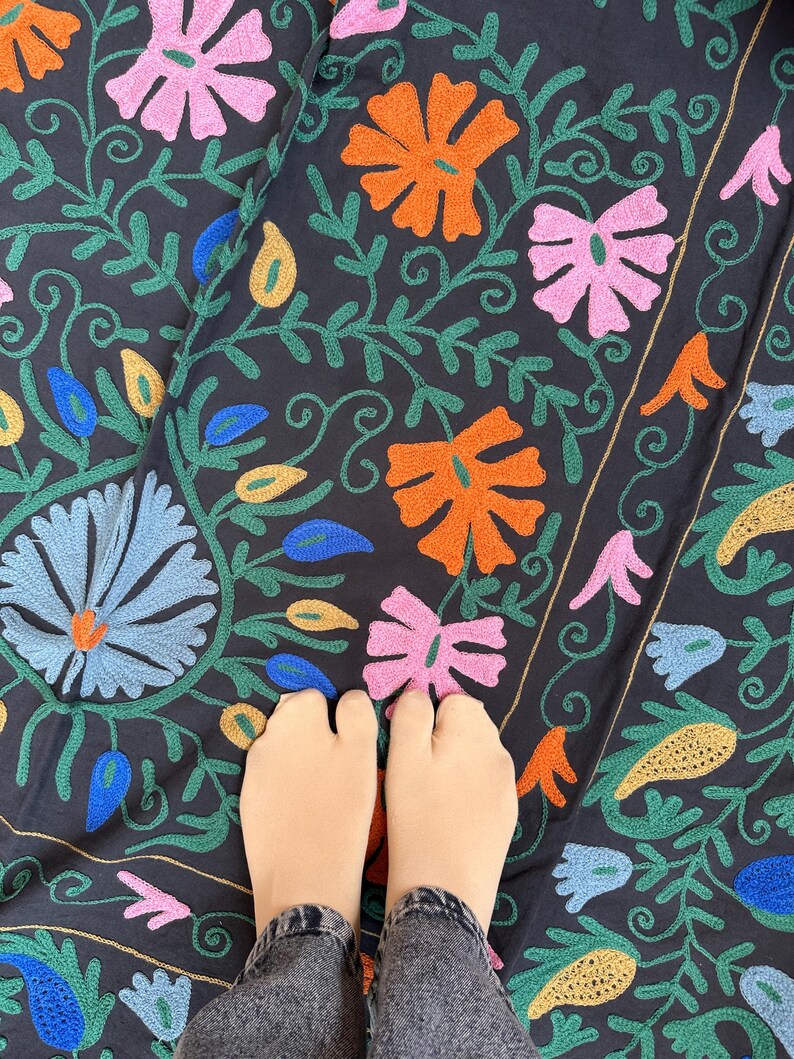 May include: Dark navy fabric with colorful floral embroidery. The design features orange, pink, blue, and yellow flowers with green vines. The embroidery is detailed and covers the fabric. The image shows a close-up of the fabric.