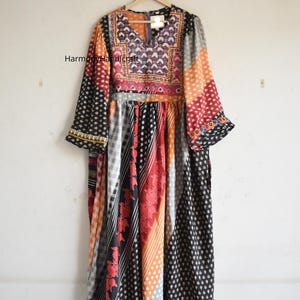 May include: A long, patchwork dress with a V-neck and long sleeves. The dress features a variety of patterns and colors, including black, red, orange, and white. The sleeves have a ruffled cuff. The dress is hanging on a wooden hanger.