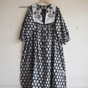 Peut inclure: Une robe à motifs noirs et blancs avec un empiècement blanc orné de motifs floraux gris et une fermeture à liens noirs. Le vêtement présente des manches trois-quarts et un imprimé géométrique répétitif. Le texte HARMONYHANDICRAFT est visible.