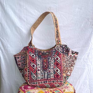 May include: A shoulder bag featuring a patchwork of embroidered fabric in shades of red, pink, white, and black. The bag displays intricate geometric patterns, small circular mirrors, and a single tan strap. The exterior is heavily textured with detailed needlework.