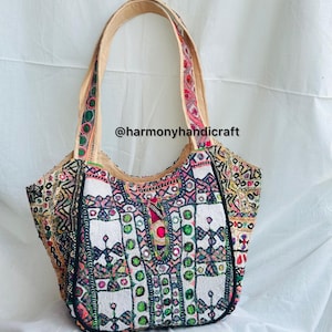 May include: A colorful tote bag with intricate embroidery and embellishments. The bag features a cream-colored handle and a white front panel with geometric patterns in pink, green, and blue. The text "@harmonyhandicraft" is visible.