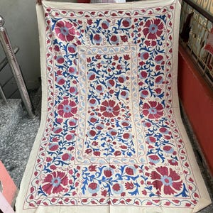 May include: A rectangular off-white textile featuring an intricate embroidered pattern of large red flowers and blue swirling vines. The design is organized with a central rectangular panel surrounded by a wide border, all showcasing floral motifs.