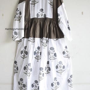 Puede incluir: Un vestido largo blanco con estampado floral gris. El vestido presenta mangas largas y un corpi&ntilde;o y paneles de hombros marrones. El vestido tiene cintura fruncida y falda fluida. El nombre de la marca "Harmony Handicraft" es visible.