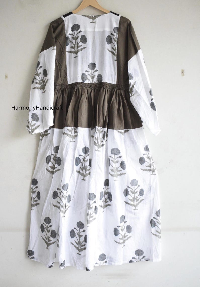 Puede incluir: Un vestido largo blanco con estampado floral gris. El vestido presenta mangas largas y un corpi&ntilde;o y paneles de hombros marrones. El vestido tiene cintura fruncida y falda fluida. El nombre de la marca "Harmony Handicraft" es visible.